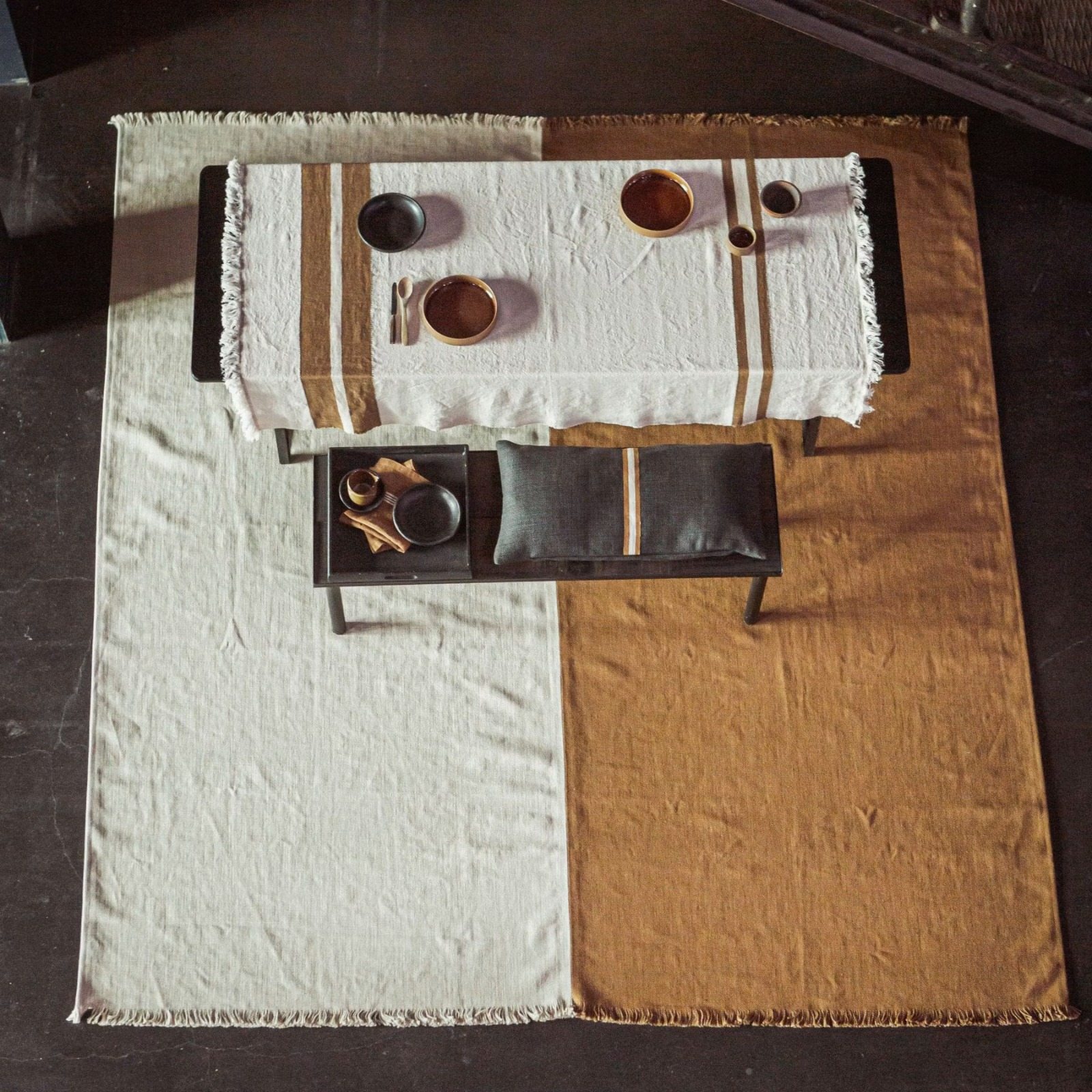 Cream linen tablecloth with ochre stripes and ceramic bowls on a table over a cream and ochre linen rug, seen from above